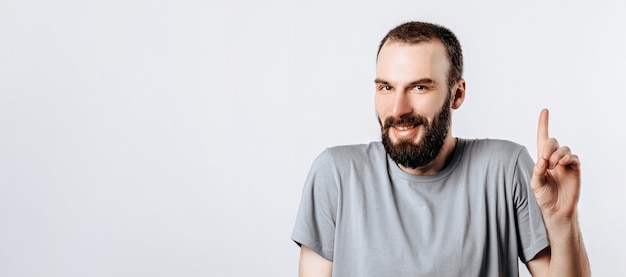 Retrato de hombre guapo joven alegre sonriendo mirando a la cámara apuntando con el dedo hacia arriba sobre fondo blanco con espacio para publicidad simulacro