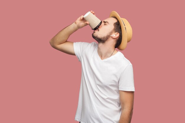 Retrato de hombre guapo joven con barba hipster en camisa blanca y sombrero casual de pie y tomando café en vaso de papel, estilo de vida de frescura. Interior, aislado, foto de estudio, fondo rosa