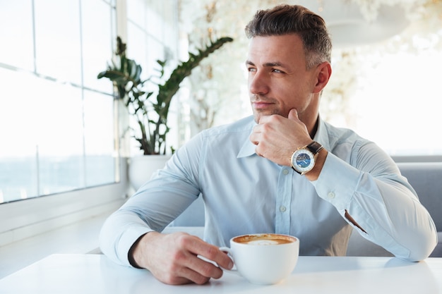 Retrato de hombre guapo sentado solo en el café de la ciudad con una taza de capuchino, mirando a un lado con melancólica vista y tocando la barbilla