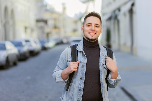 Retrato de hombre guapo sonriente en chaqueta de mezclilla y mochila en ciudad urbana