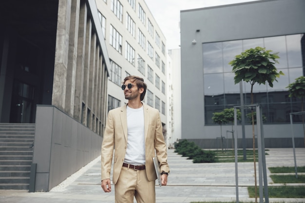 Retrato de hombre guapo sonriente en elegante traje y gafas de sol de pie cerca de un edificio moderno con portátil personal en las manos