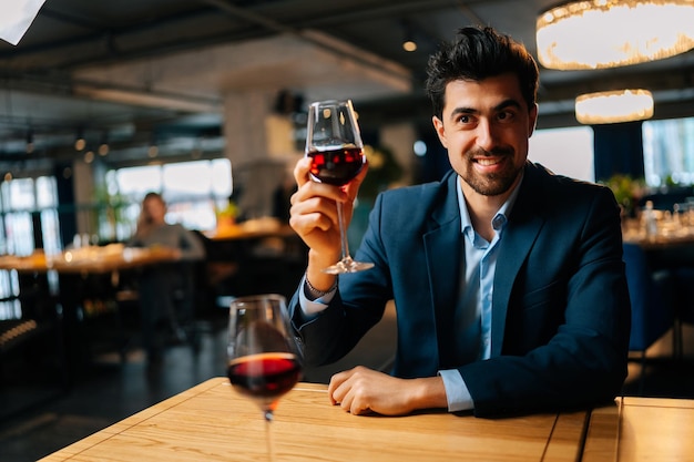 Retrato de un hombre guapo con traje elegante tintineando copas de vino tinto con una novia irreconocible mirándola sentada en la mesa en el restaurante por la noche