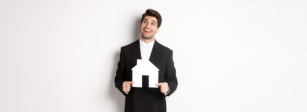 Foto retrato de un hombre guapo con traje que busca casa sosteniendo una casa de papel y mirando la esquina superior derecha c