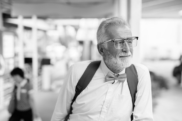 Retrato de hombre guapo turista senior barbudo con ropa elegante mientras explora la ciudad de Bangkok en blanco y negro