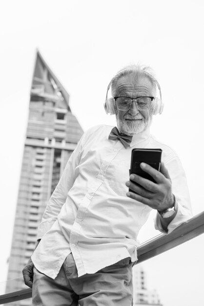 Retrato de hombre guapo turista senior barbudo con ropa elegante mientras explora la ciudad de Bangkok en blanco y negro