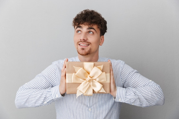 Retrato de hombre hermoso amable vestido con camisa sonriendo mientras sostiene la caja de regalo de cumpleaños aislada sobre la pared gris