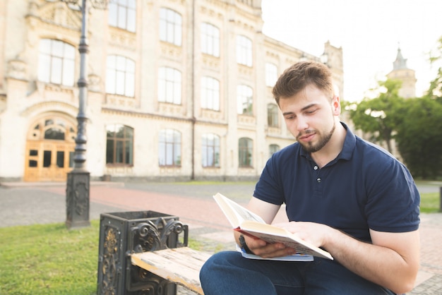 Retrato de un hombre hermoso con barba y una camiseta negra sentado en un banco en el fondo de la arquitectura y leyendo un libro.