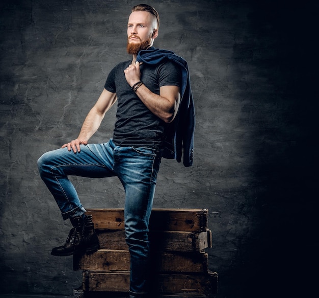 Retrato de hombre hipster barbudo pelirrojo vestido con una chaqueta azul posando en una caja de madera sobre fondo gris.