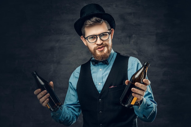 Retrato de hombre hipster barbudo vestido con una camisa azul, chaleco elegante y sombrero de copa sostiene dos botellas de cerveza artesanal con una idea de una fiesta.