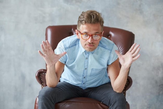 Retrato de un hombre inteligente con estilo mira fijamente a la cámara pequeña camisa azul carismática sin afeitar sentada en una silla de cuero marrón diálogo negociación manga corta peinado brutal Foto de alta calidad