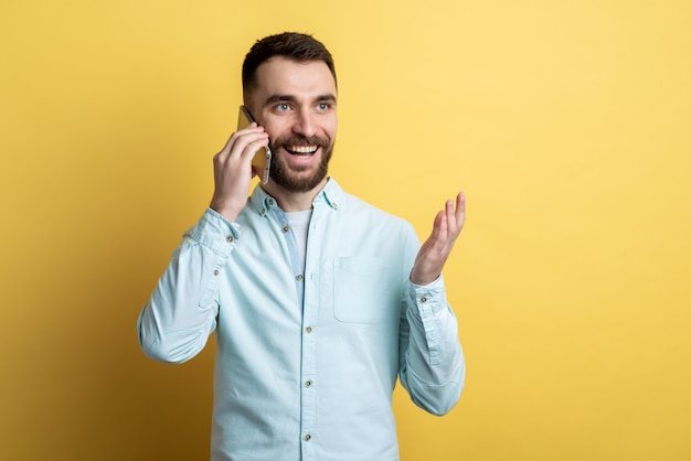 Retrato de hombre joven aislado en la pared amarilla hablando por teléfono inteligente con alegría de emoción