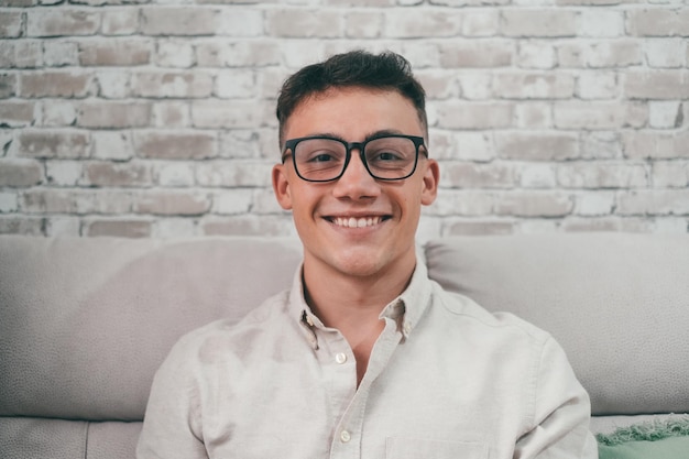 Retrato de un hombre joven y atractivo mirando a la cámara sonriendo y divirtiéndose sentado en el sofá Adolescente masculino feliz con anteojos enjoyxA