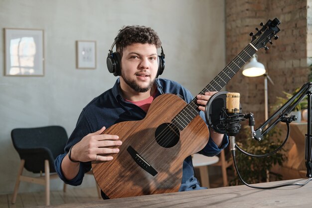 Retrato de hombre joven en auriculares mirando a la cámara y tocando la guitarra durante el aire en la radio