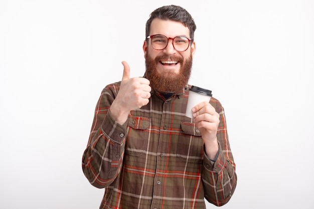 Retrato de hombre joven con barba feliz bebiendo su café de la mañana y mostrando los pulgares para arriba