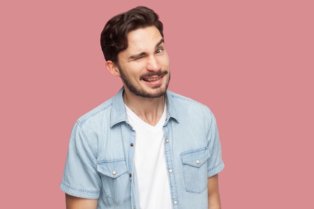 Retrato de hombre joven barbudo guapo divertido en camisa azul estilo casual de pie, guiñando un ojo y mirando a cámara con cara divertida y sonrisa con dientes. tiro del estudio de interior, aislado en fondo rosado.