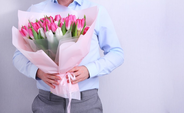 Retrato de un hombre joven en camisa azul con ramo de tulipanes rosados y blancos en gris. Tarjeta de felicitación de primavera. Pascua, concepto de flor de primavera. Día de la madre o de la mujer.