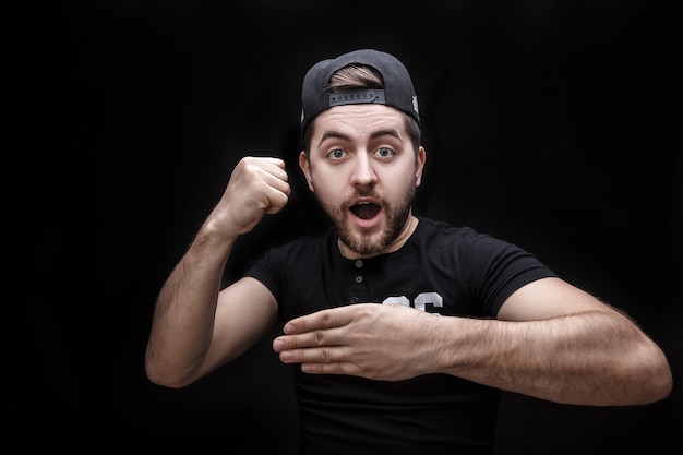 Retrato de hombre joven en camisa y gorra sobre fondo negro hace el truco de la mano