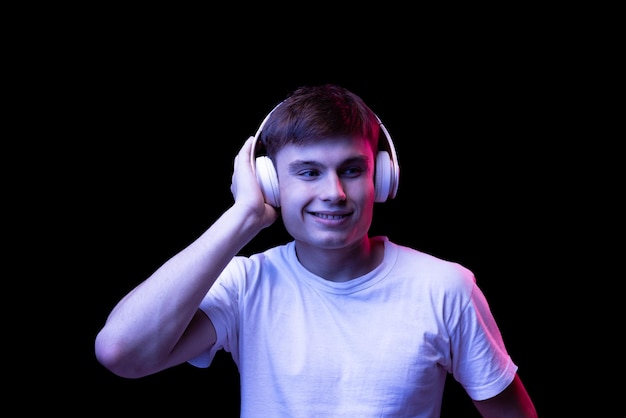 Retrato de hombre joven en camiseta blanca posando escuchando música en auriculares aislados sobre negro