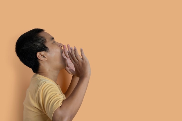 Retrato de hombre joven con camiseta casual gritando y gritando fuerte a un lado con la mano en la boca aislado en el fondo naranja del estudio Emoción humana cara expresión concepto copia espacio