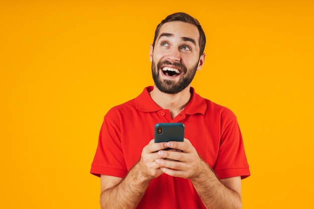 Retrato de hombre joven en camiseta roja sonriendo y sosteniendo smartphone aislado sobre amarillo