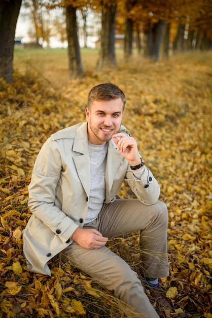 Retrato de un hombre joven en una capa en los árboles de otoño. Un hombre se sienta en el suelo.