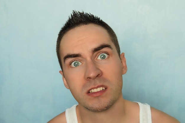 Retrato de hombre joven con expresión asustada sobre un fondo azul ojos sorprendidos Emoción sorprendida en el rostro humano chico guapo de ojos azules
