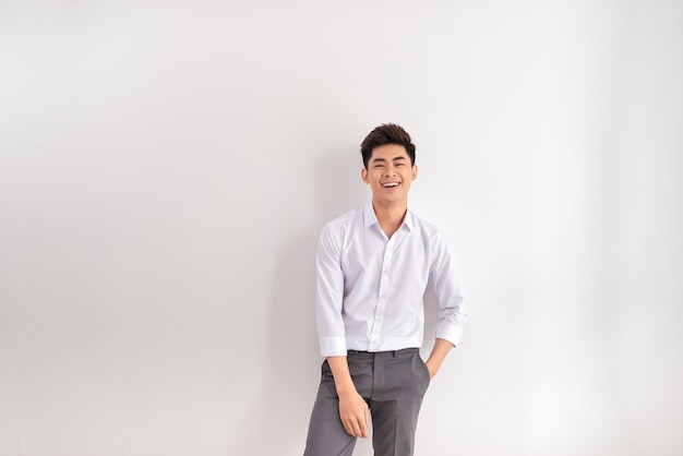 Foto retrato de hombre joven feliz de pie contra el fondo blanco. hombre asiático con las manos en el bolsillo apoyado contra la pared blanca