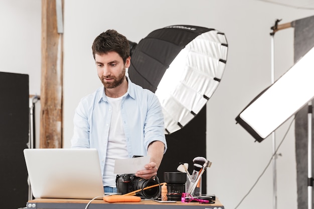 Retrato de hombre joven fotógrafo morena usando laptop mientras dispara modelo con cámara profesional y softbox en estudio