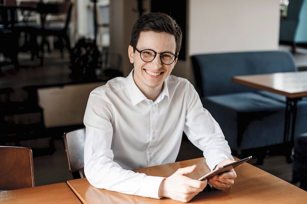 Retrato de un hombre joven guapo positivo sentado en un escritorio y mirando a la cámara sonriendo mientras sostiene una tableta con anteojos.