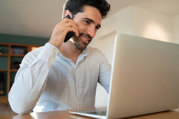 Retrato de hombre joven hablando por su teléfono móvil y trabajando desde casa con el portátil. Concepto de oficina en casa.