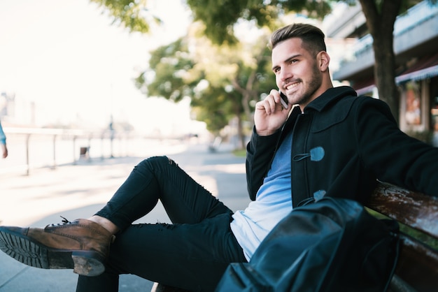 Retrato de hombre joven hablando por teléfono mientras está sentado en un banco al aire libre en la calle. Concepto de comunicación.