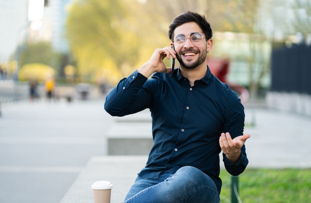 Retrato de hombre joven hablando por teléfono mientras está sentado en un banco al aire libre. Concepto urbano.