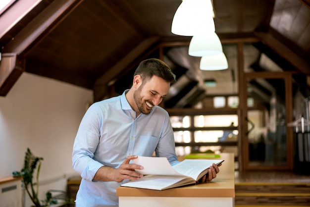 Retrato del hombre joven hermoso que se relaja en interior y hojea a través del libro.