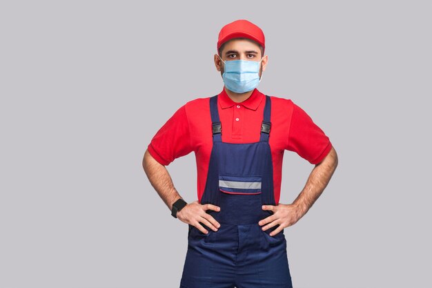 Retrato de hombre joven con máscara médica quirúrgica en general azul, camiseta roja y gorra de pie y tomados de la mano en la cintura y mirando a cámara, interior, tiro del estudio, aislado sobre fondo gris