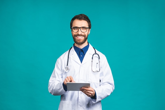 Foto retrato de hombre joven médico barbudo con estetoscopio sobre el cuello en abrigo médico de pie contra el fondo azul aislado.