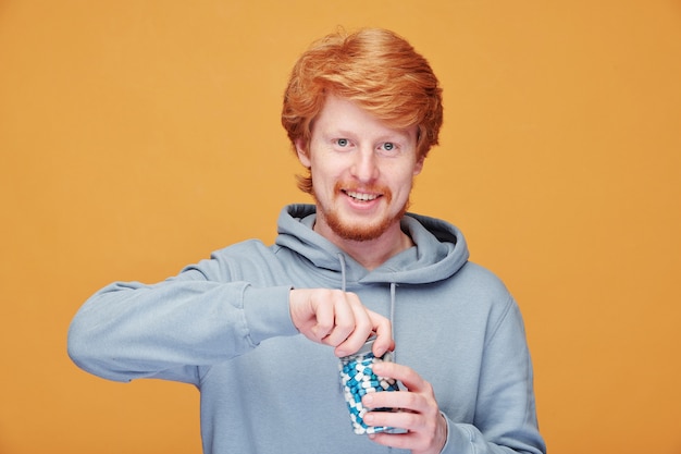 Retrato de hombre joven pelirrojo alegre con capucha abriendo botella de vitamina mientras cuida su inmunidad