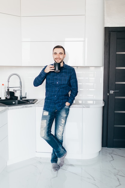 Retrato de hombre joven de pie en la cocina sosteniendo una taza de café mirando lejos en el pensamiento