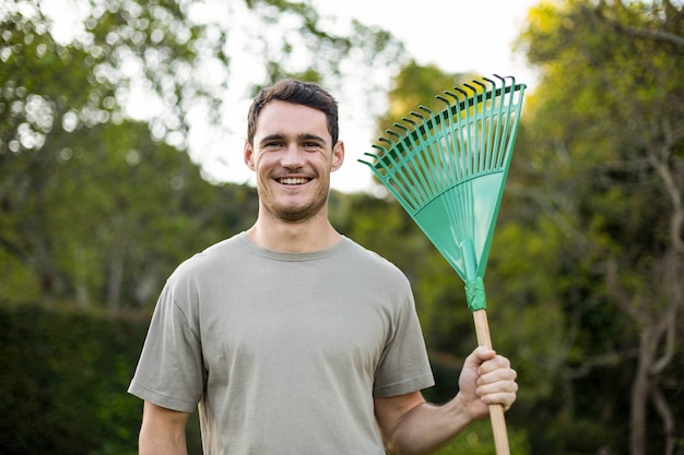 Retrato de hombre joven de pie con un rastrillo de jardinería en el jardín