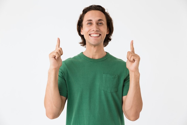 Retrato de hombre joven positivo con camiseta básica sonriendo y apuntando con el dedo hacia arriba en copyspace aislado sobre pared blanca