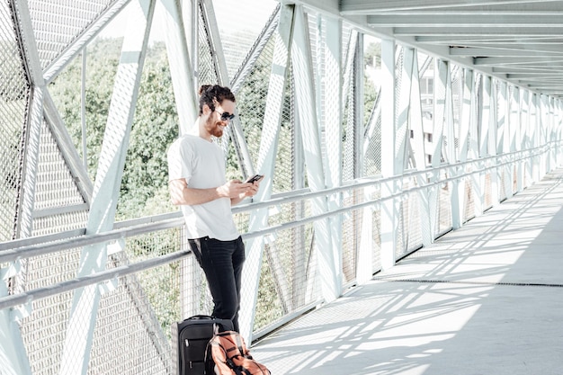 Retrato de un hombre joven en ropa casual con un teléfono en el fondo de la terminal del aeropuerto moderno con teléfono inteligente. Viajero haciendo llamada, mensajería. Estación de autobuses. Hombre hipster con equipaje. Dupdo