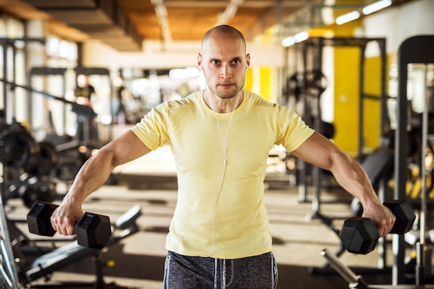 Retrato del hombre joven sano muscular activo fuerte que levanta pesas de gimnasia con los brazos abiertos y que escucha música en el gimnasio.