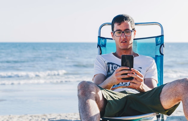 Retrato de hombre joven sentado en la hamaca mirando teléfono inteligente en la playa