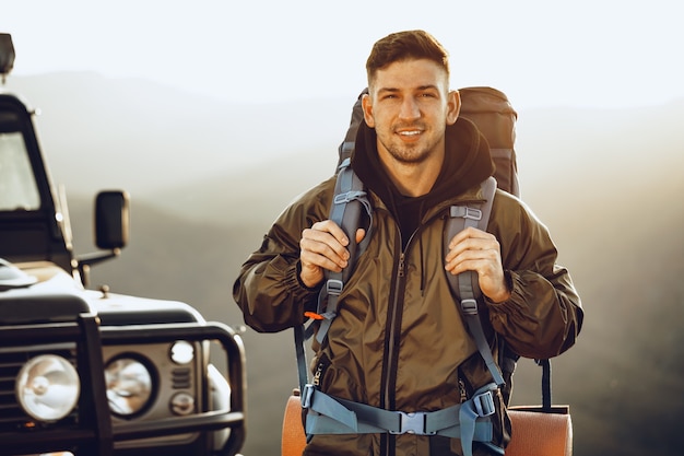 Retrato de un hombre joven viajero en equipo de senderismo de pie cerca de su coche todoterreno