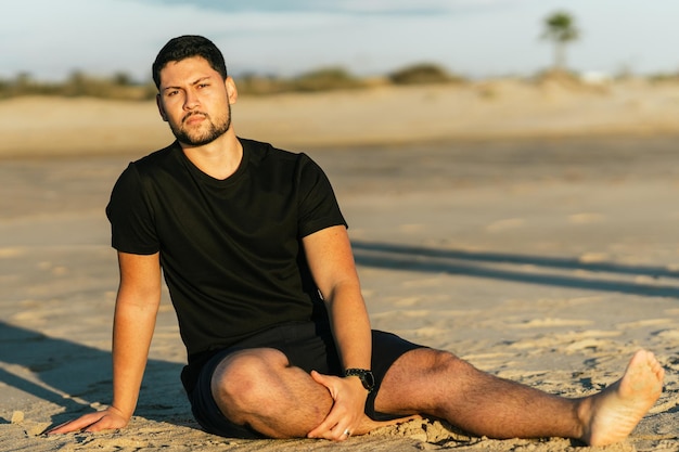 Retrato de un hombre latino fitness sentado en la arena de la playa durante el amanecer