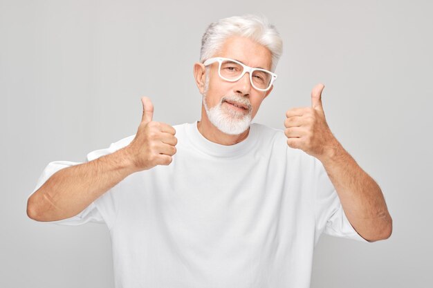 Retrato de un hombre maduro con camiseta blanca y gafas sonriendo alegremente mostrando los pulgares hacia arriba gesto aislado en el fondo blanco del estudio Aprueba una buena elección decisión correcta