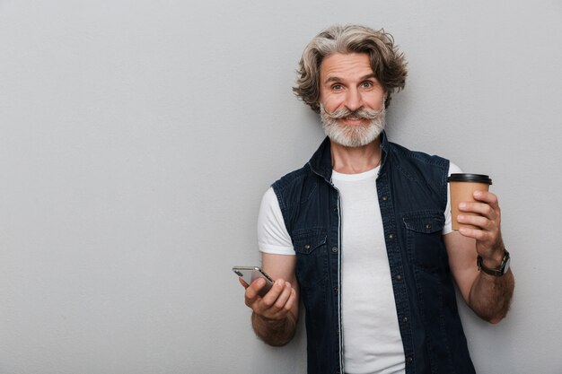 Retrato de un hombre maduro elegante sonriente hermoso que lleva un chaleco que se encuentran aisladas sobre fondo gris, utilizando el teléfono móvil mientras bebe café para llevar