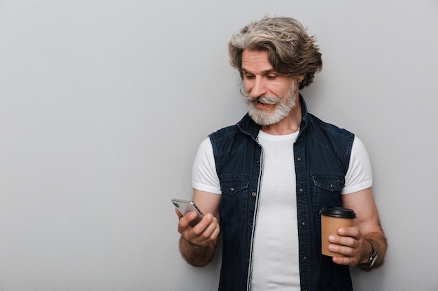 Foto retrato de un hombre maduro elegante sonriente hermoso que lleva un chaleco que se encuentran aisladas sobre fondo gris, utilizando el teléfono móvil mientras bebe café para llevar