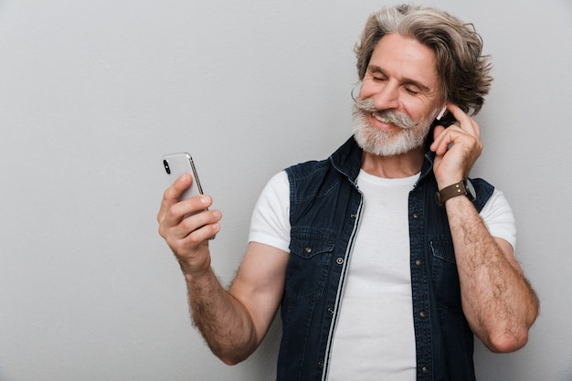 Retrato de un hombre maduro con estilo guapo sonriente con un chaleco que se encuentran aisladas sobre fondo gris, escuchando música con auriculares, sosteniendo el teléfono móvil