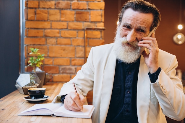 Retrato de un hombre mayor barbudo tomando café y usando el teléfono inteligente en la cafetería. Retrato del hombre barbudo gris feliz que se sienta en café.