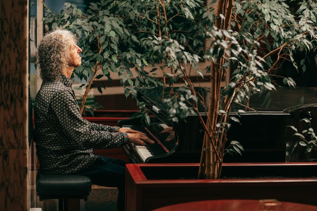 Retrato de hombre mayor canoso con cabello largo y rizado está sentado al piano y tocando música, disfrutando de una agradable velada en el hotel. Foto de estilo de vida